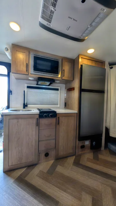 Bright rv rental kitchen showing a modern sink, stovetop, microwave, and large refrigerator within light wood cabinetry.. Ember Overland 2023