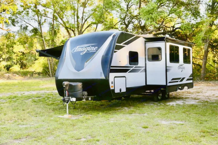 A white & black Grand Design Imagine RV is parked on a grassy campsite with trees, ready for your next RV rental adventure.. Grand Design Imagine 2020
