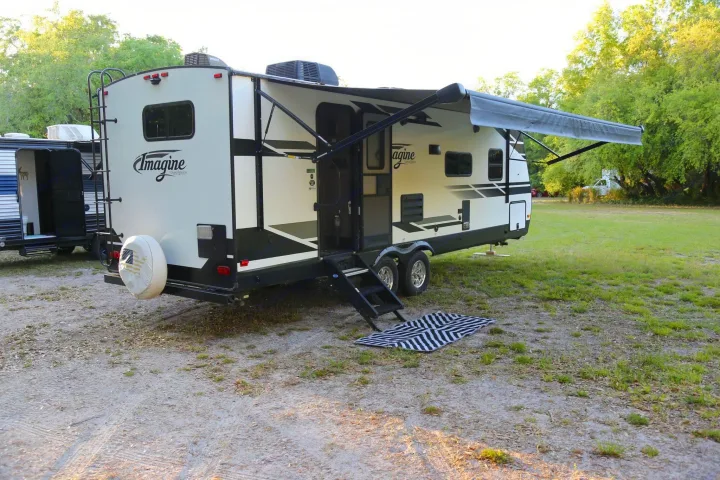 This white RV rental, a travel trailer, is parked at a campsite with its awning extended, steps out, and a striped mat.. Grand Design Imagine 2020
