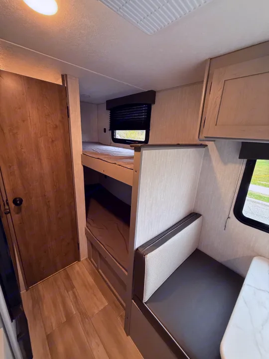 RV rental interior: light wood-look floor, bunk beds with gray bedding, a gray and light dinette, and a window with blind.. Coachmen Clipper 17CBH 2025