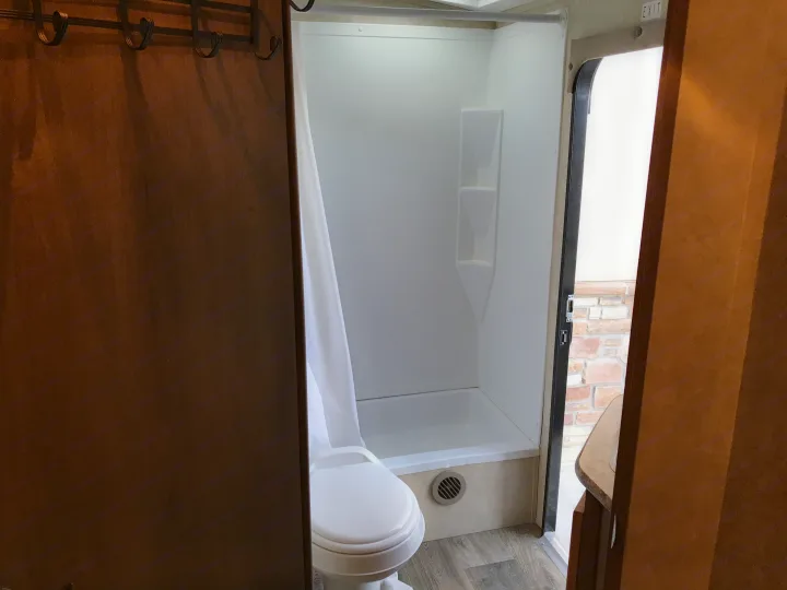 RV rental bathroom: compact design with a white shower, toilet, and shower curtain, visible with a view of a brick wall outside.. Cruiser RV Shadow Cruiser 2015