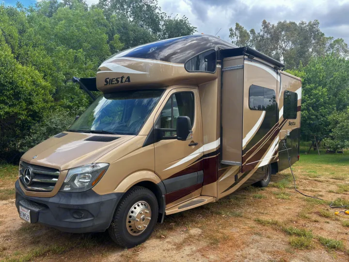 A beige & brown Mercedes-Benz Class C RV rental, with its slide-out extended, parked outdoors on a dirt patch.. Thor Siesta 2016