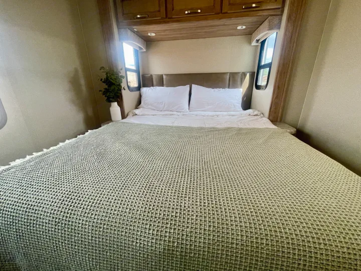 An RV rental bedroom featuring a full bed with white pillows and a light green waffle-knit blanket.. Entegra Coach Vision 31V 2019