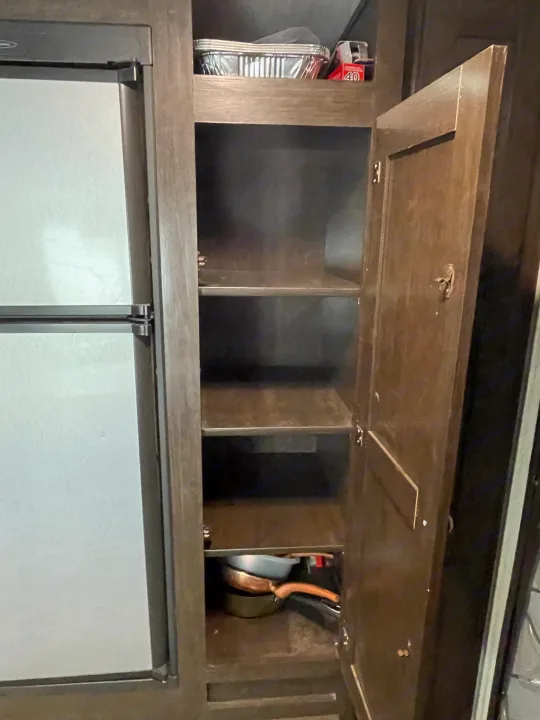 This RV rental offers practical kitchen storage: an open pantry cabinet with shelves, holding pans and foil containers, beside a refrigerator.. Heartland RVs Trail Runner 2019