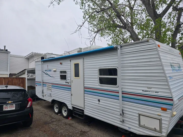This white Fleetwood travel trailer, ideal for rv rental adventures, is parked outdoors beside a black car on a cloudy day.. Fleetwood Mallard 2004