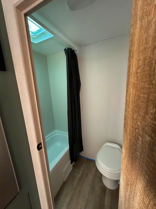 Compact RV rental bathroom: features a white tub/shower with a black curtain, toilet, and overhead skylight.. Heartland Eddie Bauer 2025