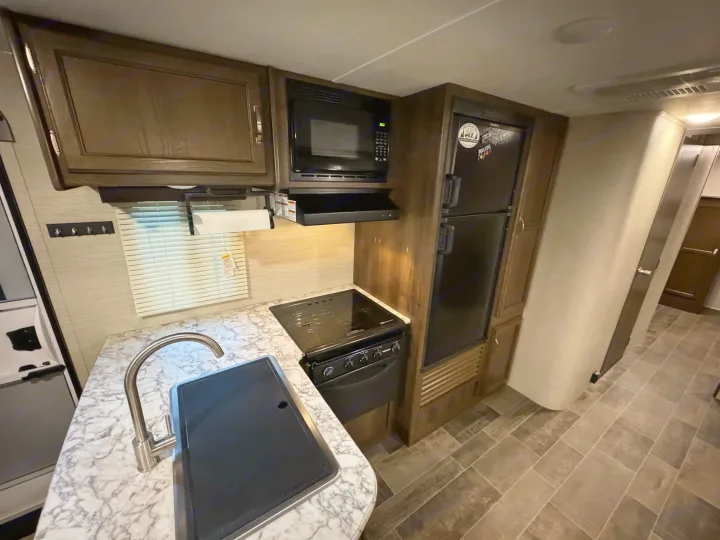 Compact RV rental kitchen with a modern sink, countertop, oven, microwave, fridge, and cabinets. Ready for travel!. Keystone RV Bullet Ultra Lite 2019