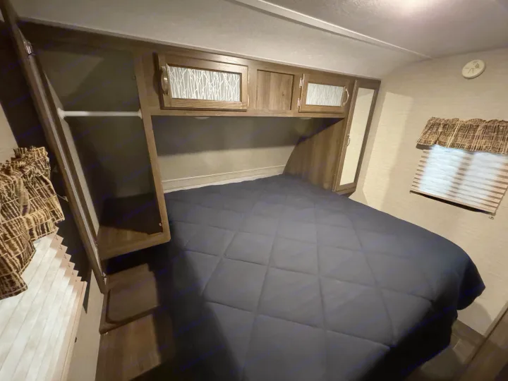 Experience comfort in this RV rental bedroom, featuring a dark bed, compact overhead cabinets with decorative glass, and a wardrobe.. Keystone RV Bullet Ultra Lite 2019
