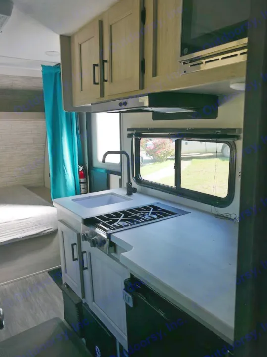 RV rental interior: Equipped kitchen with sink, stove, microwave, cabinets. Features a window view and a bed with blue curtain.. Forest River Aurora 2024