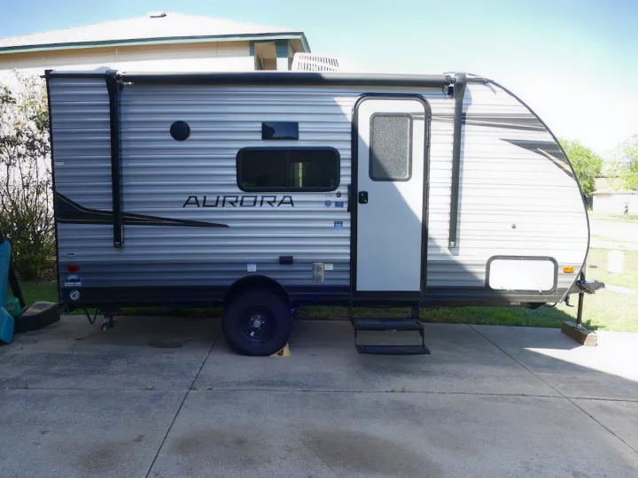 A parked gray and black "Aurora" RV rental travel trailer with an awning and steps on a concrete driveway.. Forest River Aurora 2024