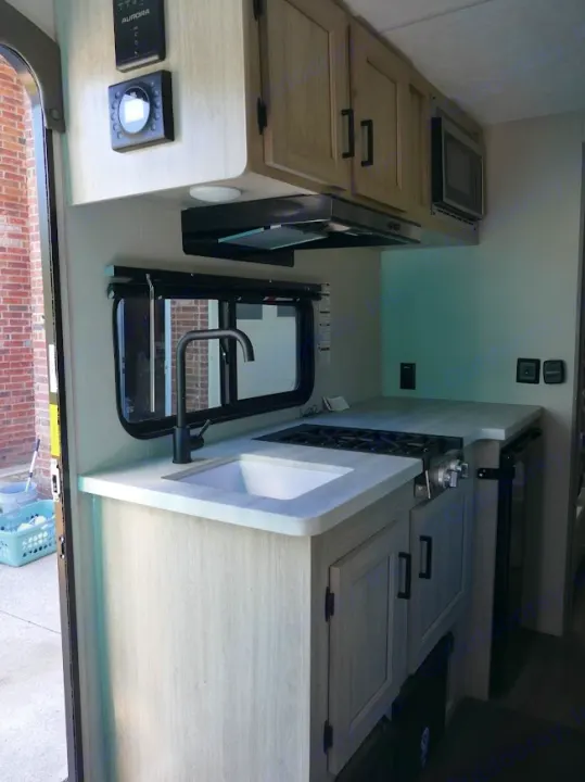 Modern RV rental kitchen interior: sink, two-burner stove, microwave, light wood cabinets, black fixtures.. Forest River Aurora 2024