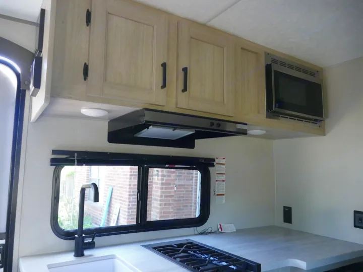 Modern RV rental interior with light wood kitchen cabinets, microwave, gas stove, sink, and range hood over the counter.. Forest River Aurora 2024