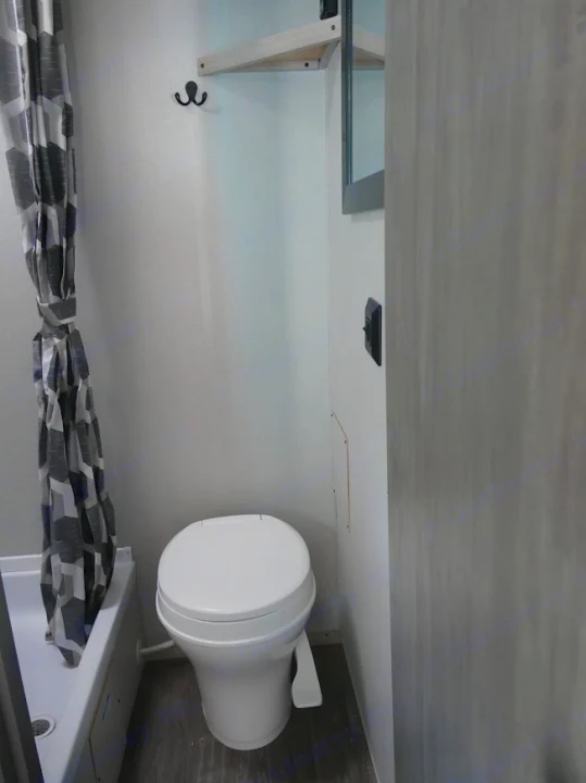 RV rental bathroom: A white toilet, grey geometric shower curtain, and small shower. Light walls, wood floor, and storage shelf.. Forest River Aurora 2024