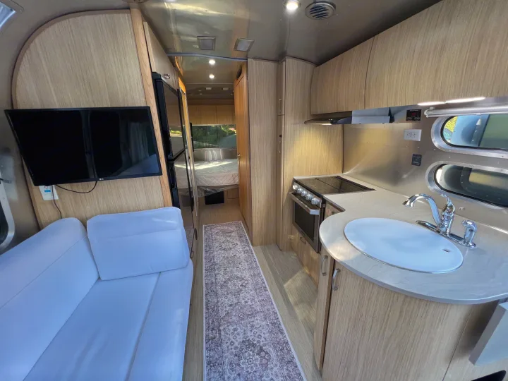 Modern rv rental interior: kitchen with stove, living area with sofa & TV, and a cozy bedroom. Ready for travel.. Airstream Flying Cloud