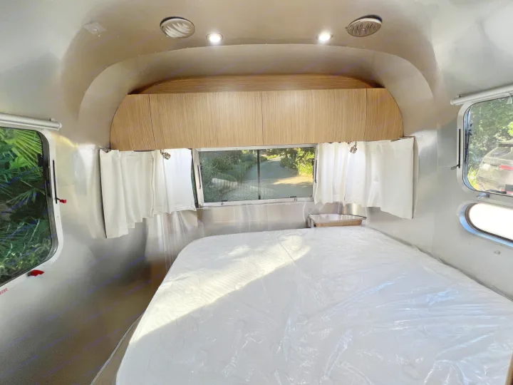 Airstream RV rental bedroom with a clean, plastic-wrapped mattress, wood cabinets, and windows with white curtains.. Airstream Flying Cloud