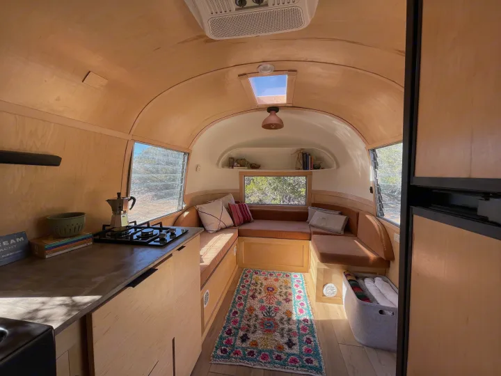 Bright rv rental interior: light wood paneling, compact kitchen, and comfy lounge area with colorful rug.. Airstream Safari 1964
