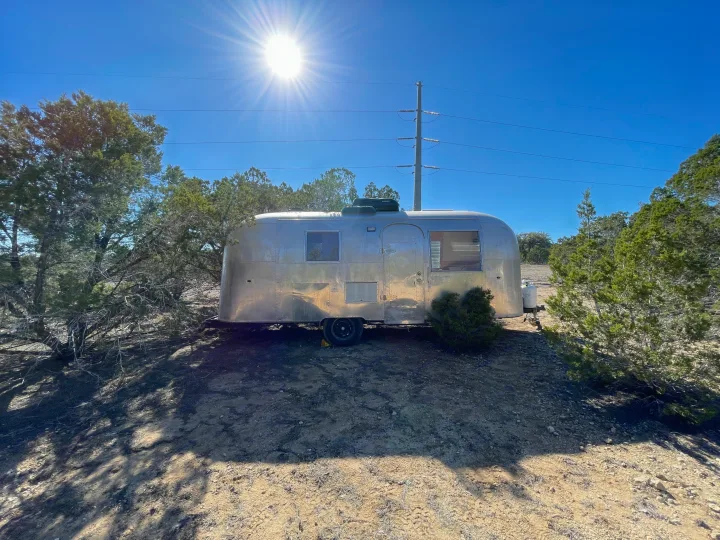 A classic silver Airstream RV rental is ready for your escape, parked in a sunny, arid outdoor setting among sparse trees.. Airstream Safari 1964