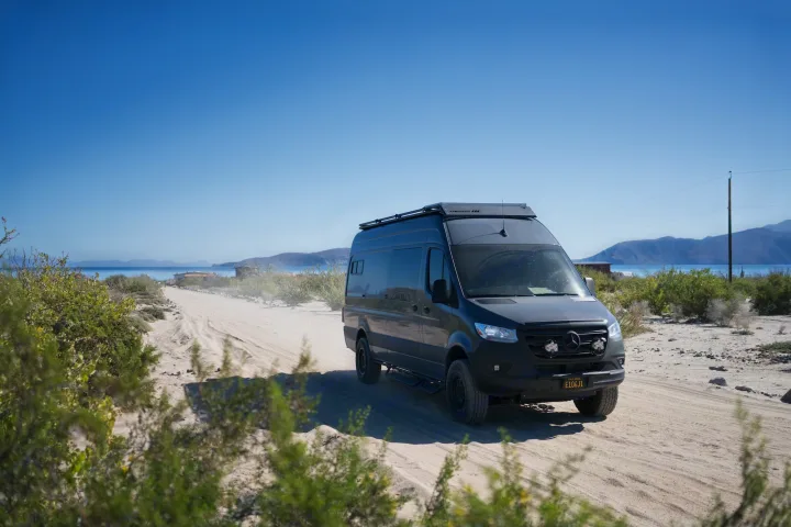 An RV rental, a dark Sprinter van, drives on a sandy coastal road, kicking up dust under a clear blue sky. Mountains in distance.. Mercedes-Benz Sprinter 2022