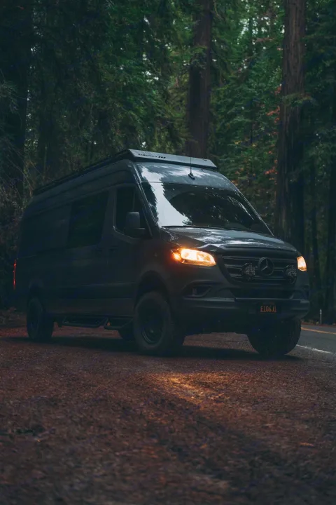A dark Mercedes Sprinter camper van, ready for adventure or rv rental, illuminates a deep forest road at dusk.. Mercedes-Benz Sprinter 2022