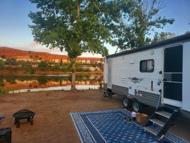 RV rental parked at a serene lakeside campsite. A blue outdoor rug and fire pit create a comfortable outdoor living space.. Forest River Shasta 2021