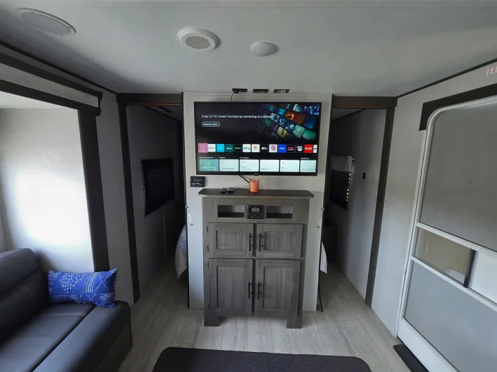 View of an RV rental living area featuring a modern entertainment center with a smart TV, sofa, and access to two rear rooms.. Forest River Shasta 2021
