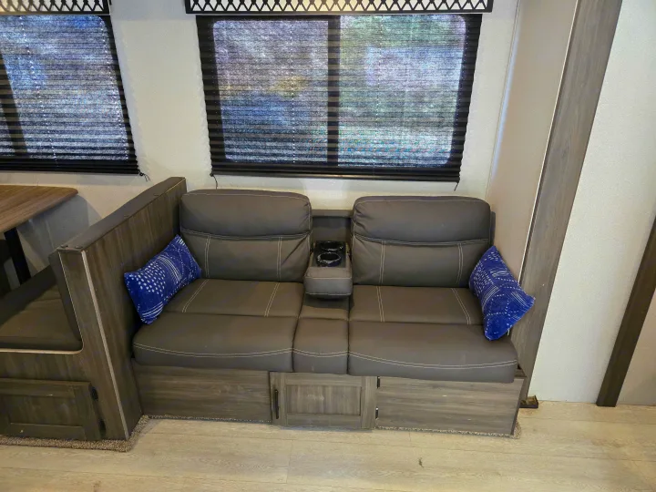 Comfortable RV rental interior featuring a gray sofa with cup holders, blue pillows, and pleated window blinds.. Forest River Shasta 2021