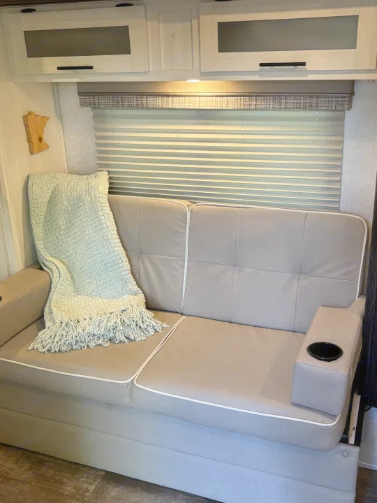 Inside an RV rental, a comfy gray sofa with a fringed blue blanket, built-in cup holder, and light wood cabinets.. Forest River R-Pod 2022
