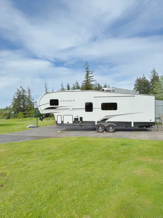 White Cougar fifth-wheel RV rental parked at a serene campsite with green grass and tall trees under a partly cloudy sky.. Keystone Cougar 2025