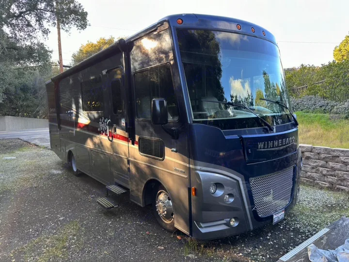 A dark blue Winnebago RV, an ideal rv rental, parked on a gravel lot. Sun highlights its side with steps extended.. Winnebago Adventurer 2021