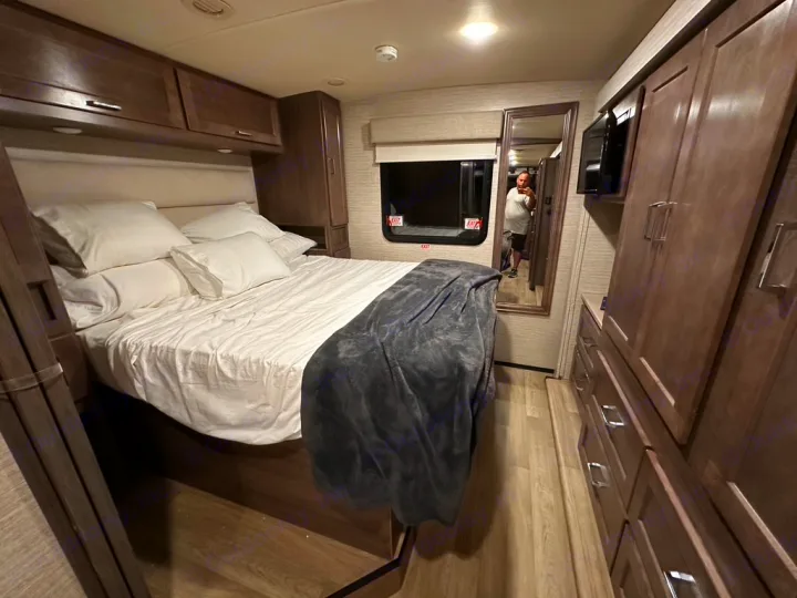 Cozy RV rental bedroom with queen bed, white bedding, built-in storage, mirror, and TV. Ready for travel.. Winnebago Adventurer 2021