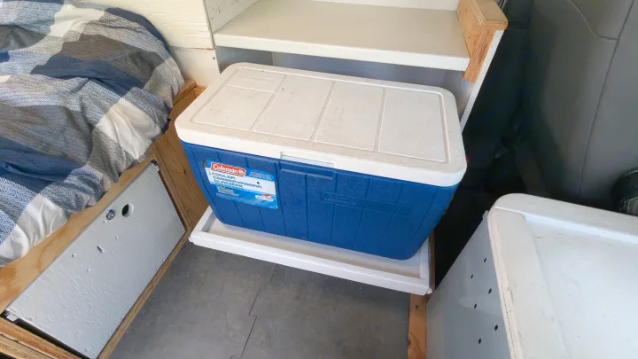 Blue Coleman cooler on a sliding tray inside an RV rental, next to a bed with a plaid blanket.. Gmc Savana 2011