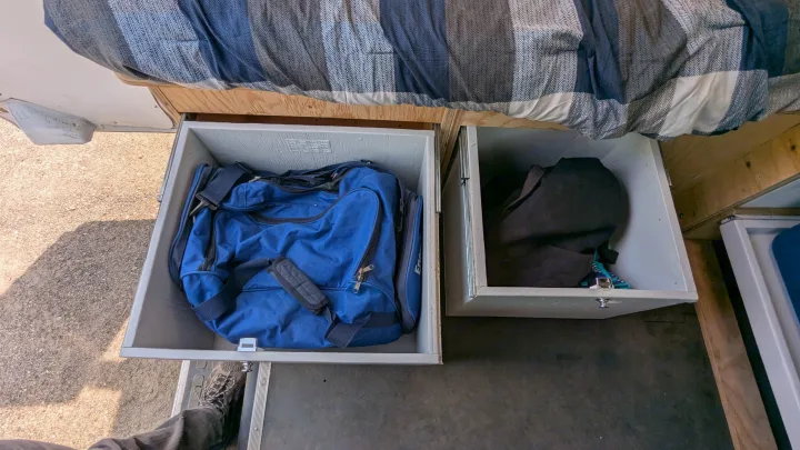 RV rental storage: Two pull-out drawers under a bed, one with a blue duffel bag, other with dark clothes in a camper van.. Gmc Savana 2011