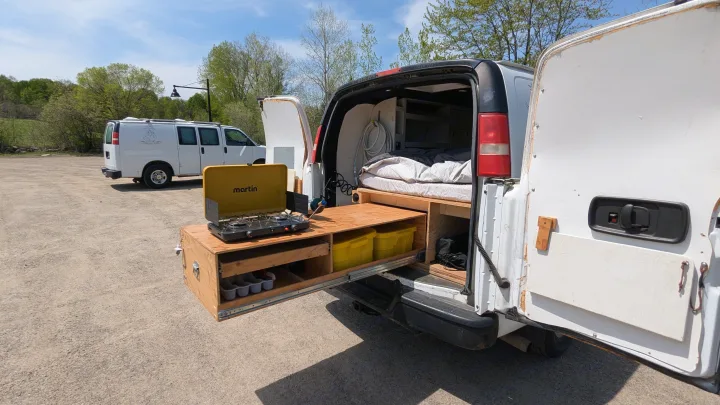 White camper RV rental van with open rear, revealing a built-in bed and a pull-out kitchen with a Martin stove for camping.. Gmc Savana 2011