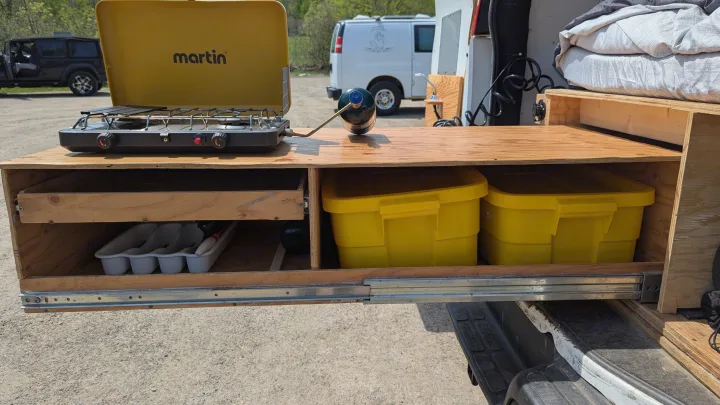 RV rental kitchen setup: A pull-out wooden counter with a Martin camping stove, propane tank, and storage bins for van life cooking.. Gmc Savana 2011
