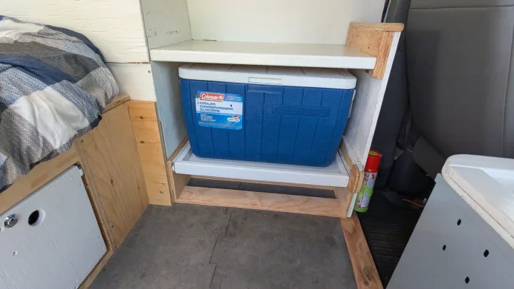 RV rental interior: blue Coleman cooler on a slide-out tray, beneath a bed with blue plaid bedding.. Gmc Savana 2011