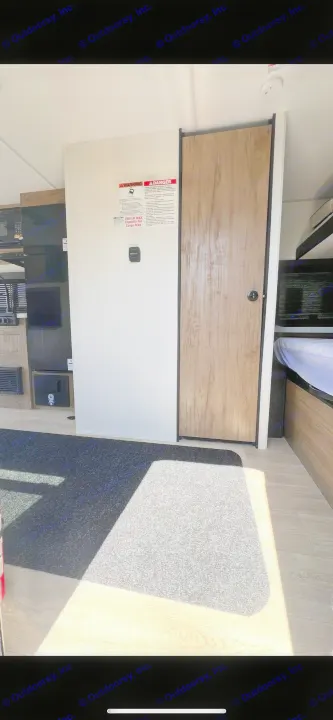 RV rental interior: View of cargo area wall with warning labels, wooden door, and part of a bed. Carpeted floor with sunlight.. Forest River Wolf Pup 2025