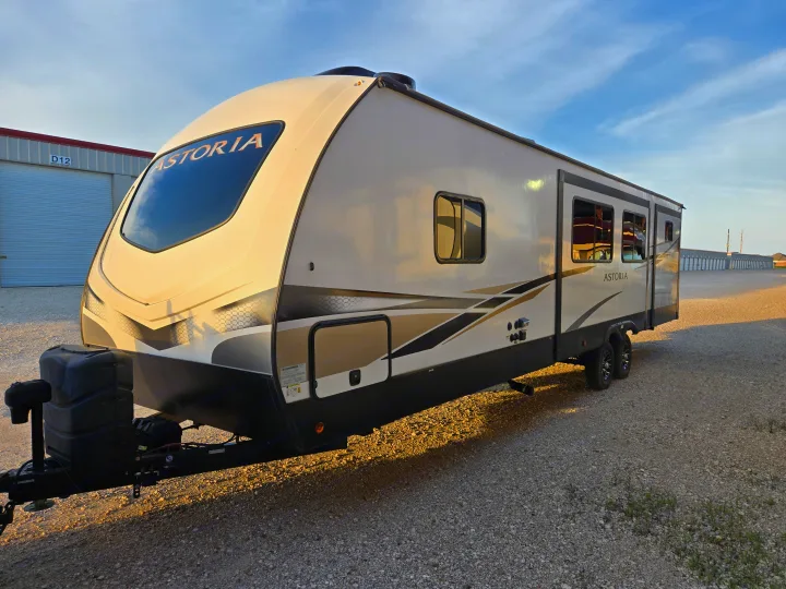 This Astoria travel trailer, ready for your next RV rental adventure, is parked on a gravel lot with storage units in the background.. Dutchman Astroia 2022