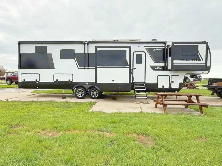 A sleek black & white EastWest Ahara fifth-wheel RV parked on a concrete pad at a campground with a picnic table. Ideal rv rental.. East to West Ahara 2024