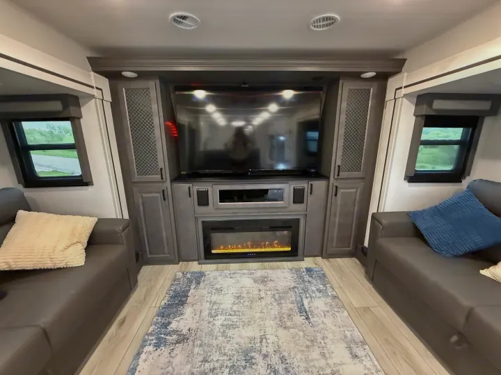 A modern RV rental interior features a large TV above an electric fireplace, flanked by two gray leather sofas with windows.. East to West Ahara 2024