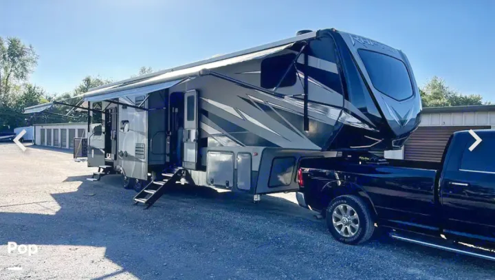 Large, dark fifth-wheel RV rental, awnings extended, hitched to a black truck on a gravel lot.. Keystone RV Raptor Toy Hauler 2020