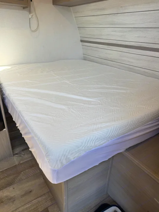 RV rental bed: White mattress cover on a modern bed with light wood interior paneling.. Dutchmen Coleman 17B 2024