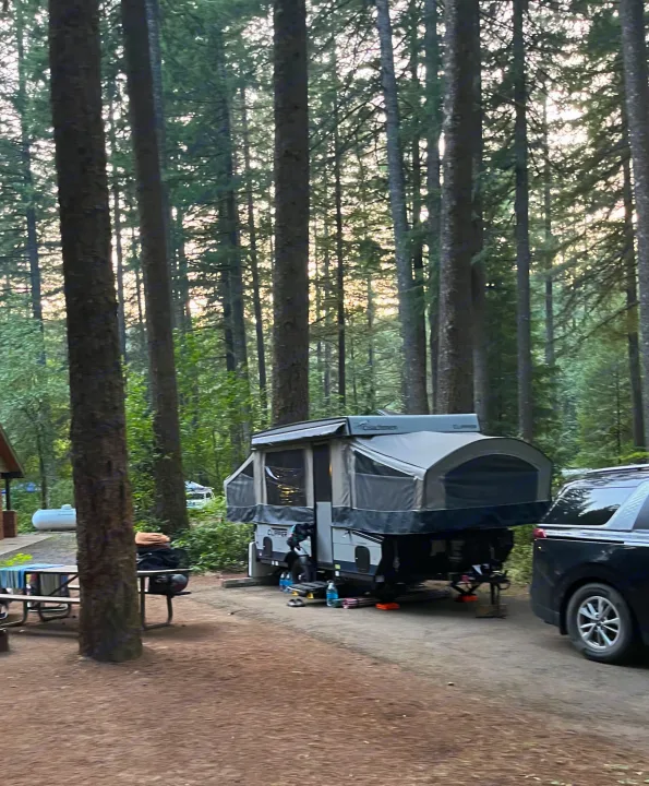 An RV rental pop-up camper is set up at a shaded forest campsite, with a parked vehicle beside it, perfect for nature getaways.. Coachmen Clipper Sport 108ST 2022