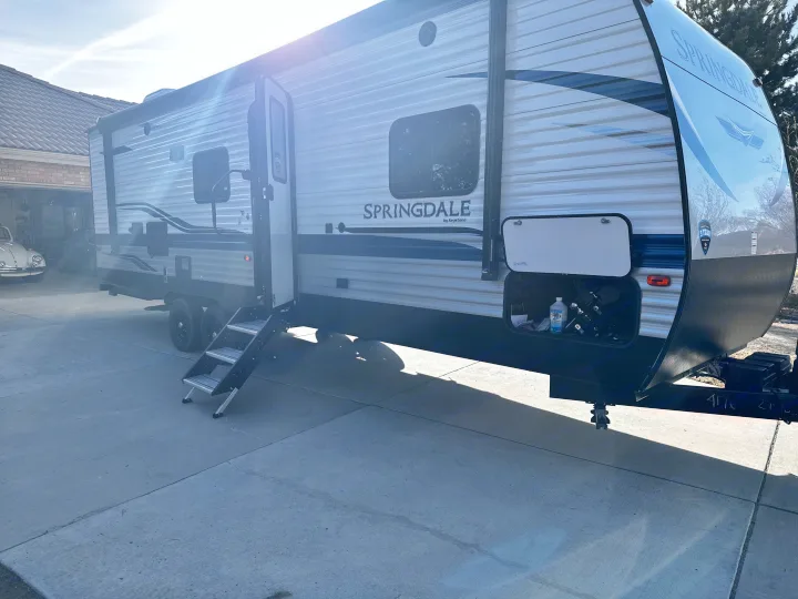 A large Springdale travel trailer (RV rental) on a sunny driveway, with its door open, steps extended, and storage compartment open.. Keystone RV Springdale 2021