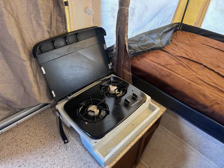 Interior of an RV rental pop-up camper, featuring a compact kitchen with a gas stove and nearby bed.. Palomino Other 2017