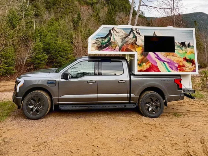 A gray F-150 Lightning electric truck with a custom, art-painted truck camper, like an rv rental, on a dirt road.. Ford Lightning 2024