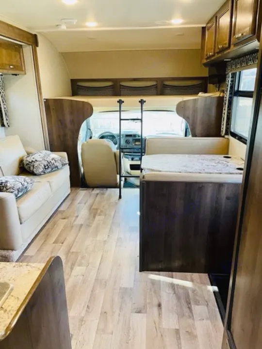 Inside a modern RV rental: light wood floors, a cozy sofa, dining space, and over-cab bunk for sleeping.. Jayco Redhawk 2018