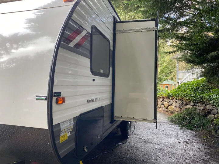 Side view of an RV rental travel trailer with an extended slide-out, parked on a wet driveway surrounded by trees.. Forest River Wildwood Sport 2021