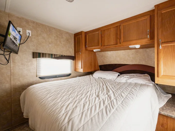 Cozy RV rental bedroom featuring a bed with white bedding, wall-mounted TV, and wooden storage cabinets.. Jayco Jay Flight 2010