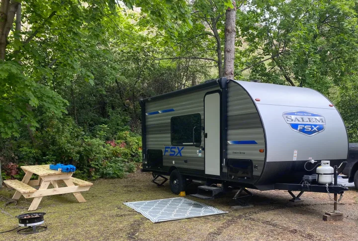 An RV rental, a compact travel trailer, is parked at a lush green campsite with a wooden picnic table and a grill.. Forest River Salem 179 2021