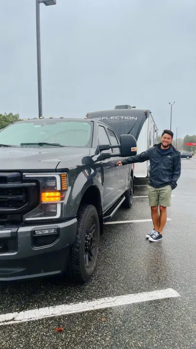 Man smiles by his truck towing a Reflection RV rental, ready for a road trip adventure on a wet day.. Grand Design Reflection 2021